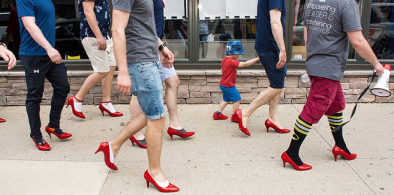 Group of men walking while wearing red high heels. Picture taken from side view.
