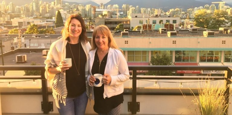 Colleen is pictured standing in front of a cityscape, beside another woman. Both are holding coffee mugs and smiling.
