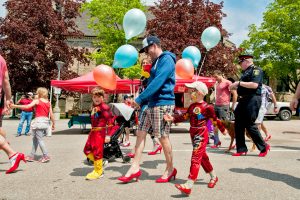 Two young boys in super hero costumes walking with a man wearing the red high heels.