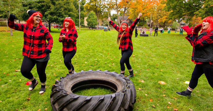 People in red plaid and plaid wigs dance around a large tractor tire