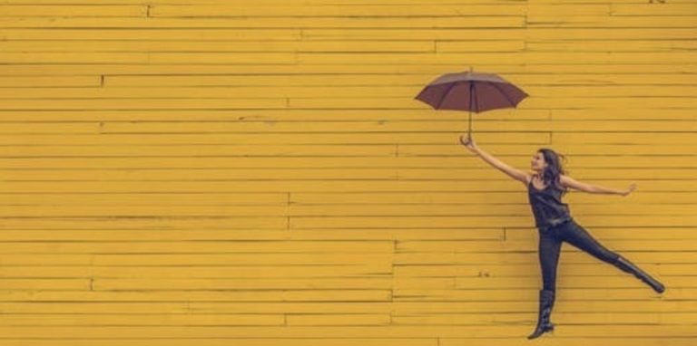 Woman holding an umbrella in front of yellow brick wall, leaping in the air.