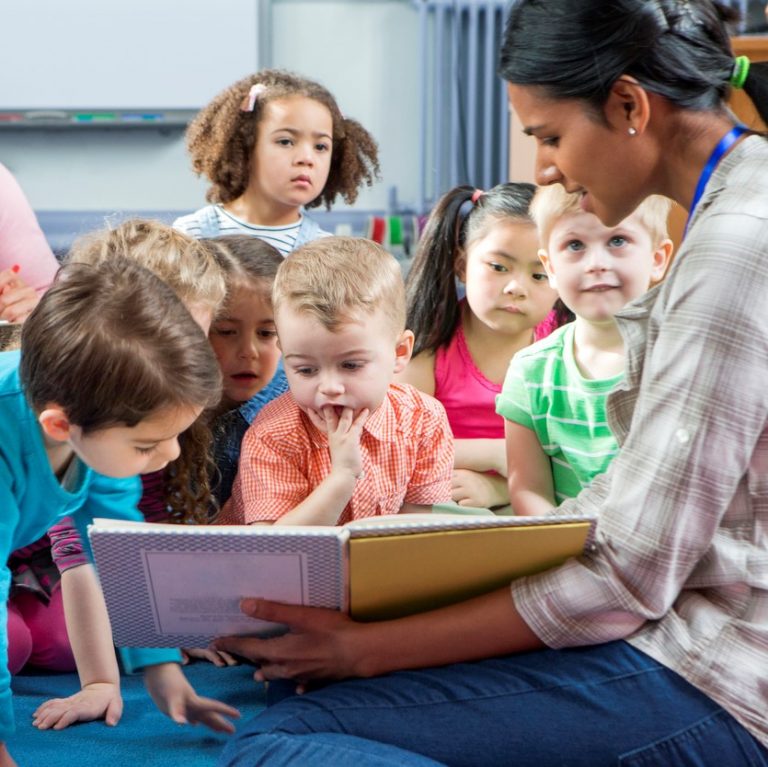 Female teacher giving a lesson to nursery students. They are sitting on the floor and there is a teacher taking notes.