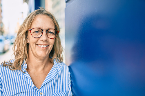 woman with glasses leans on blue wall looking at camera, She is wearing glasses, a blue bouse and has blonde hair.