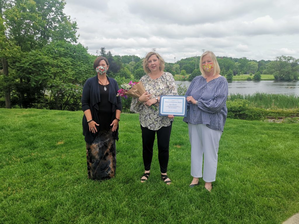 April stands on a lawn and there is a body of water behind her. On her left is YWCA Cambridge CEO, Kim Decker and on her right is YWCA Cambridge board member, Sarah Daly