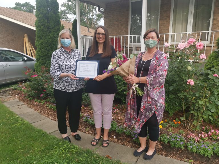 Nicole stands in her front garden holding her WOD certificate. On her right is YWCA Cambridge CEO, Kim Decker and on her left is Sarah Daly, YWCA Cambridge board memebr.