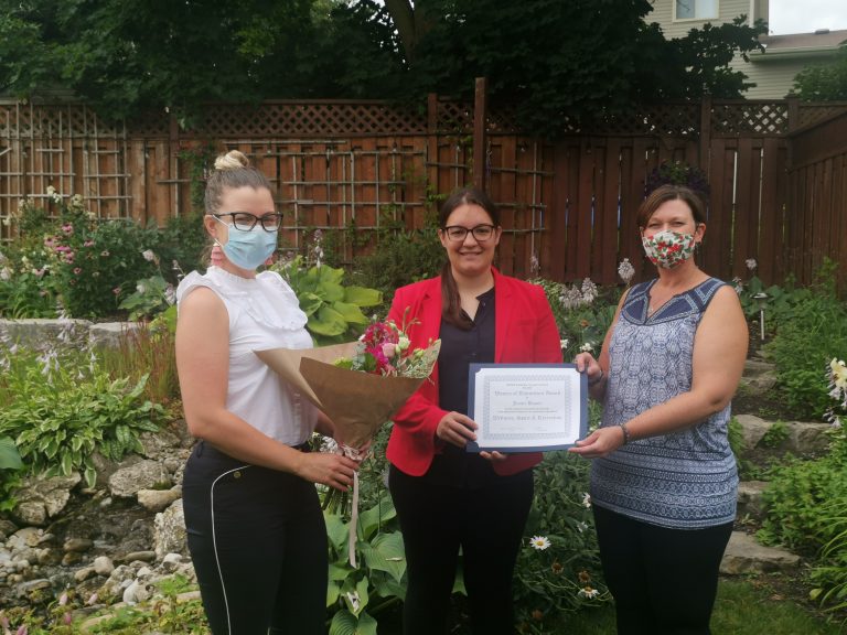 Women standing in garden holding certificate and flowers.