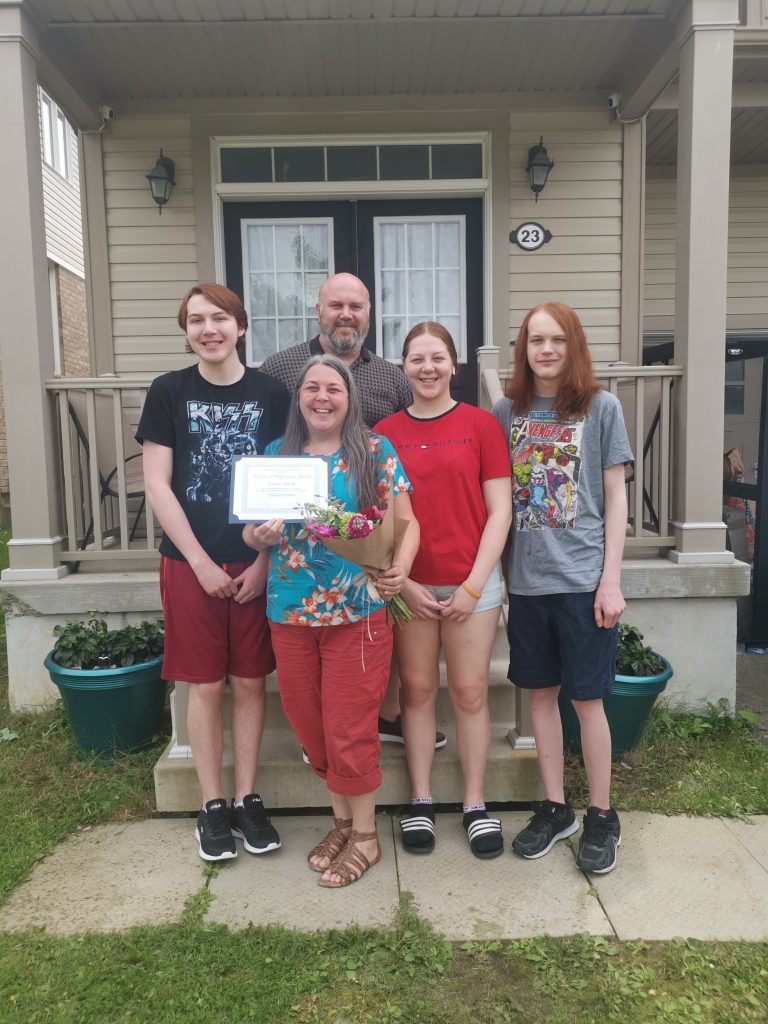 Dianne stands on her front porch holding her WOD certificate and she is surrounded by her family - her husband behind her and her 3 children on either side of her.