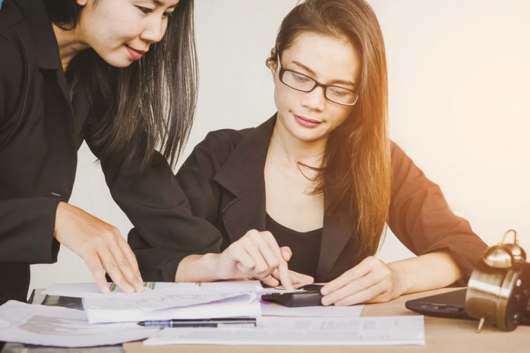 One seated woman at table with standing woman learning over her shoulder as they both review documents in a workplace setting.