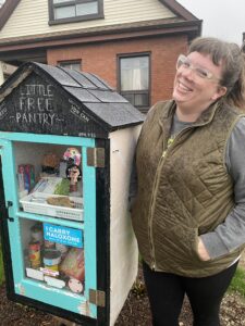 Woman standing in front of small house shaped structure with sign "little free pantry".