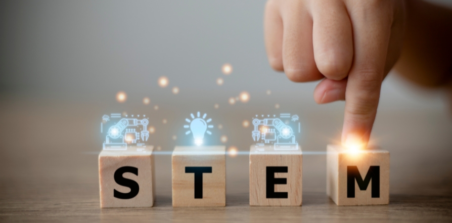 Building blocks that spell out the word "STEM" with blue icons above. Icons are of machines and a lightbulb. Childs finger is pressing down on the "M"