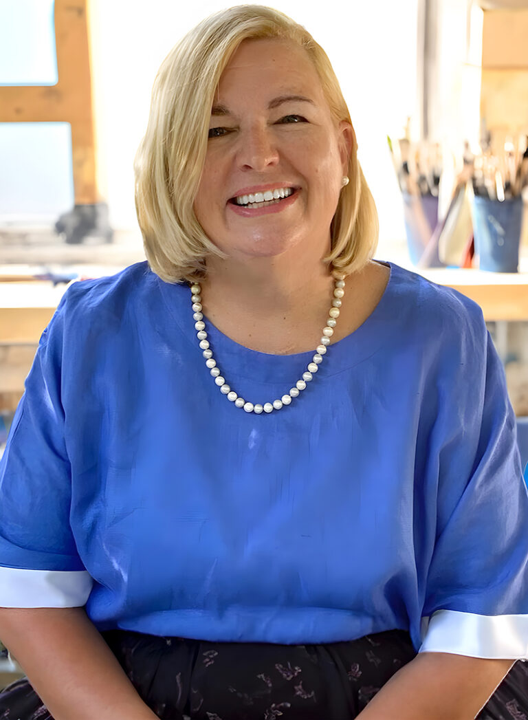 Helen wears a blue top and a pearl necklace. She's smiling and you can see from the background that she is sitting in a paint studio