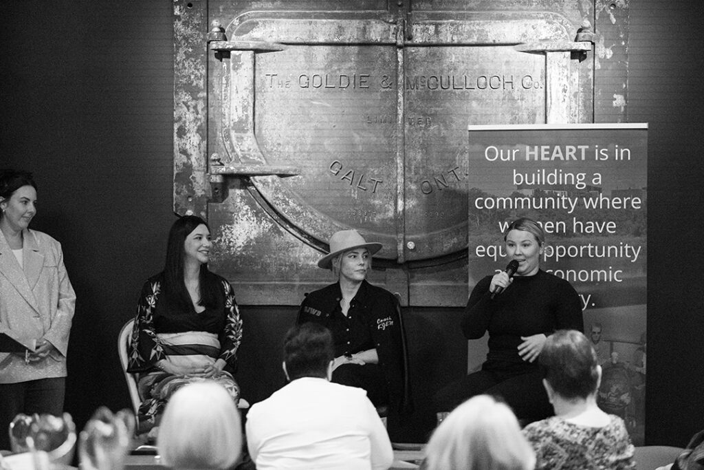 A black and white photo of She Talks speakers Kris Ejem, Mallorry Green and Sera-Lys McArthur all sitting together with MC Nataleigh Ballantyne standing beside them