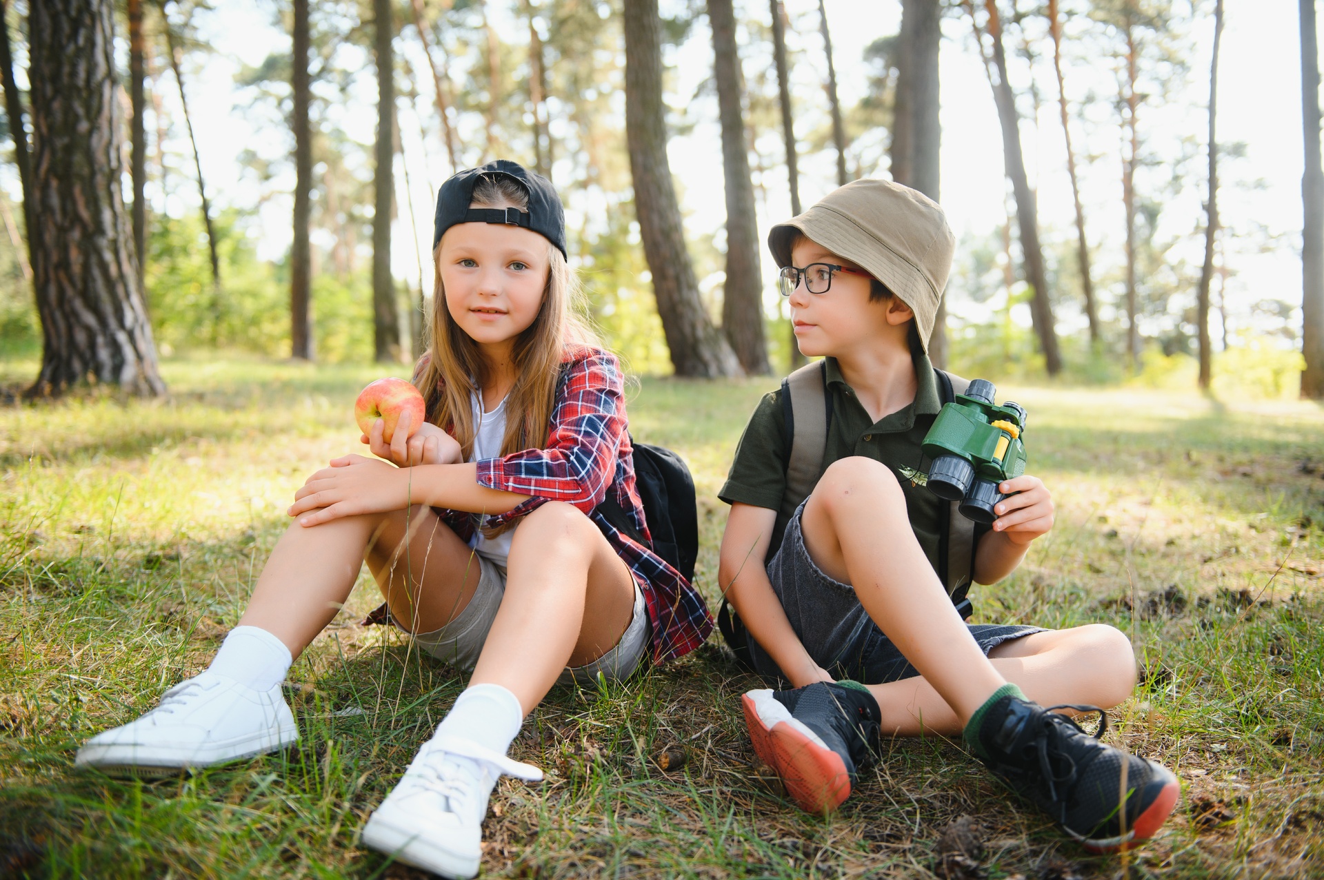 A girl and boy dressed for the outdoors sit on the grass, a forest nehind them