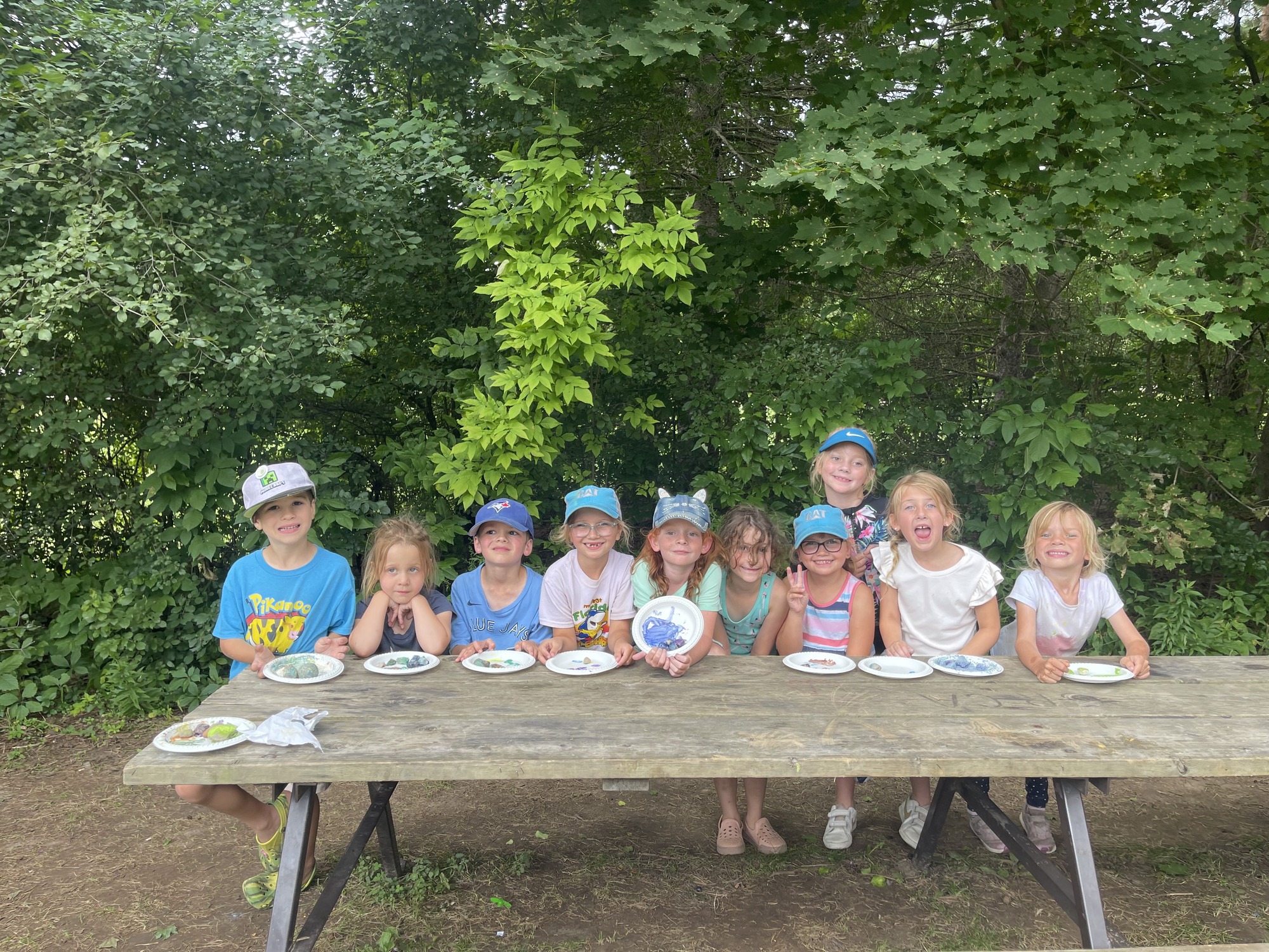 A group of about 10 wee summer campers all sitting on one side of a picnic bench, smiling and making faces for the camera