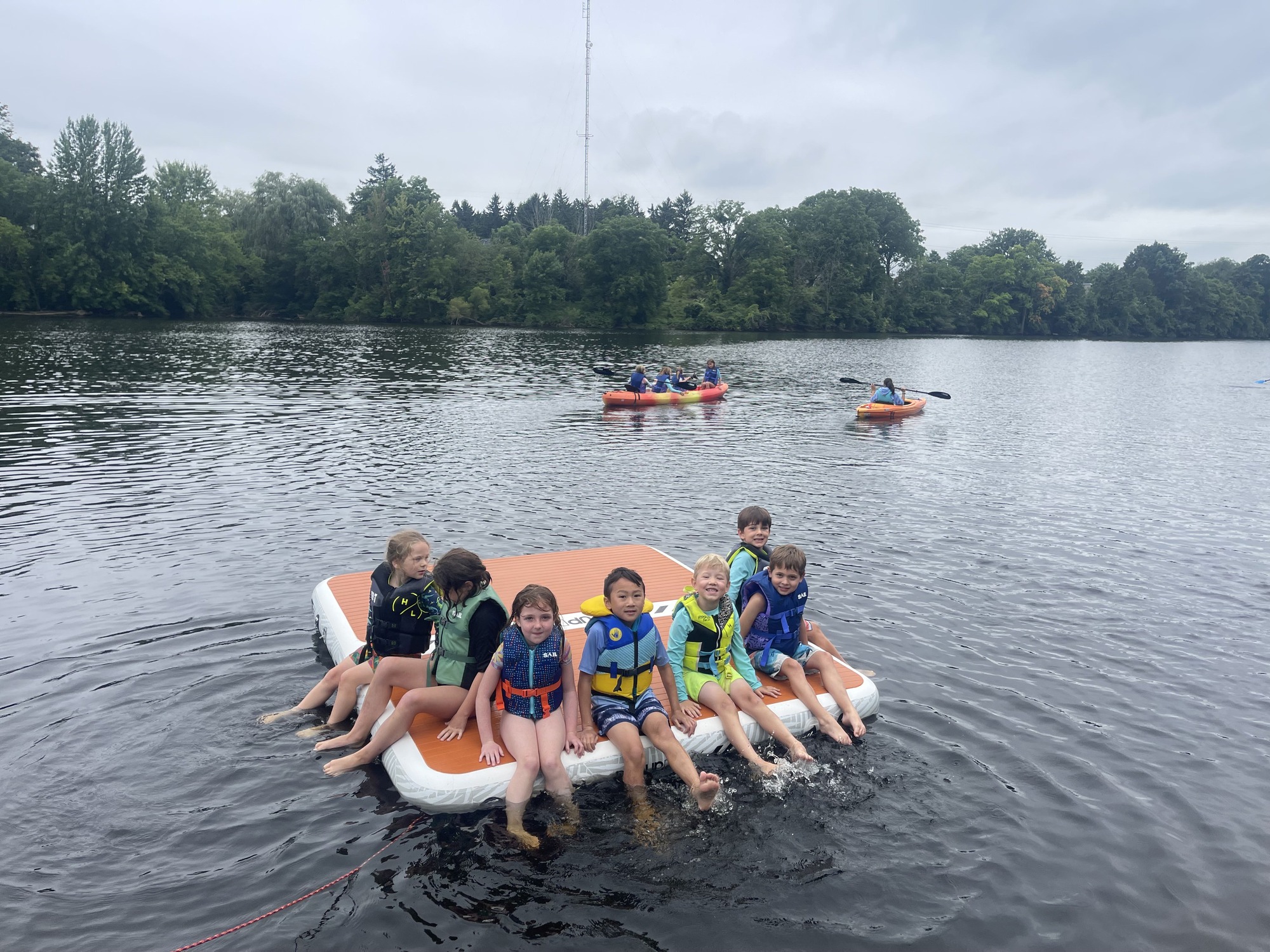 A group of campers wearing life jackets while sitting on a giant orange floatation device in the water