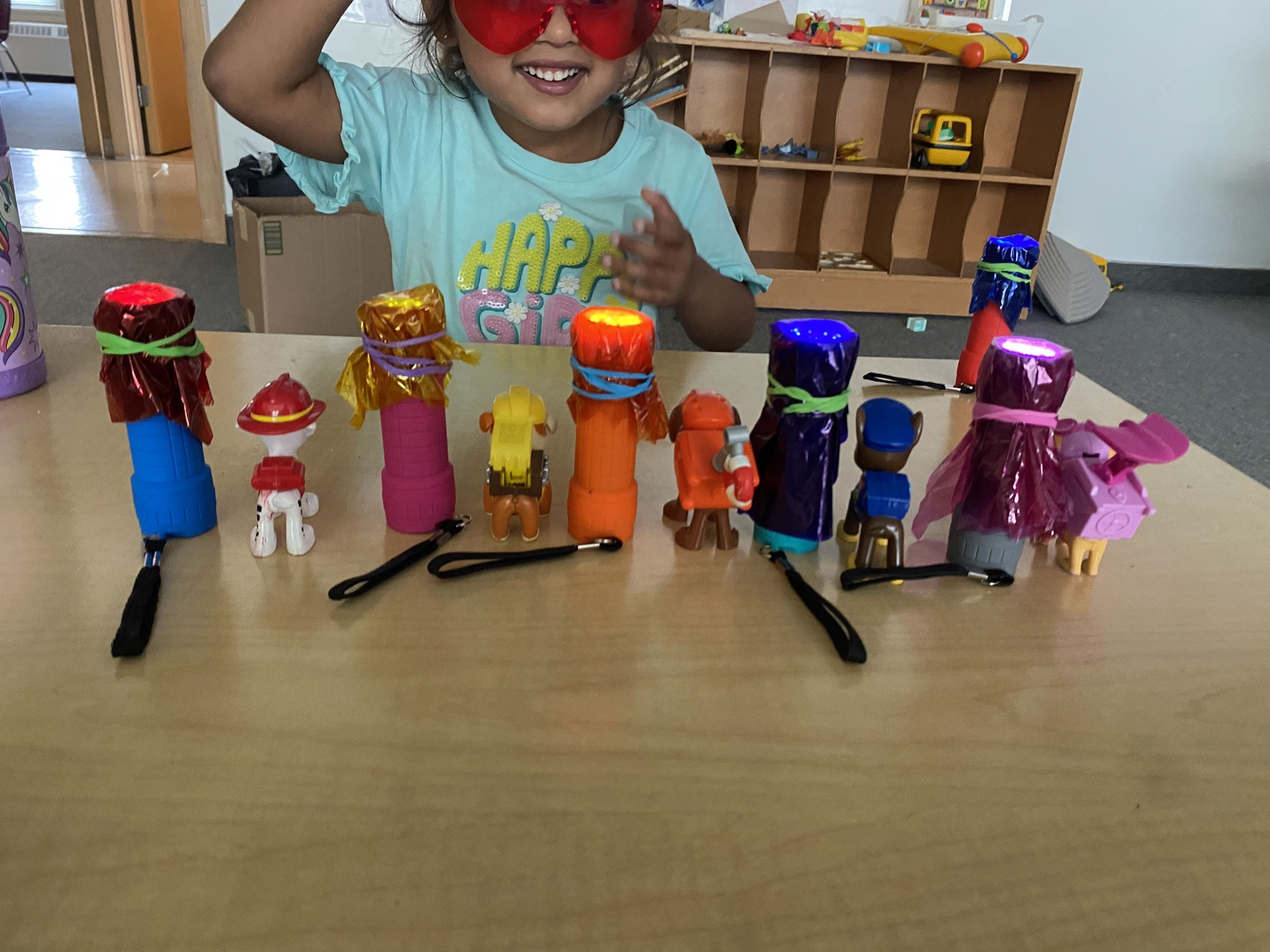 A little girl wearing a pale green tshirt and red large glasses smiles while a bunch of toys sit on a table in front of her