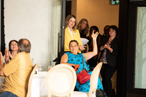 three people sit at a gala table and a group of suited women watch from an open door frame